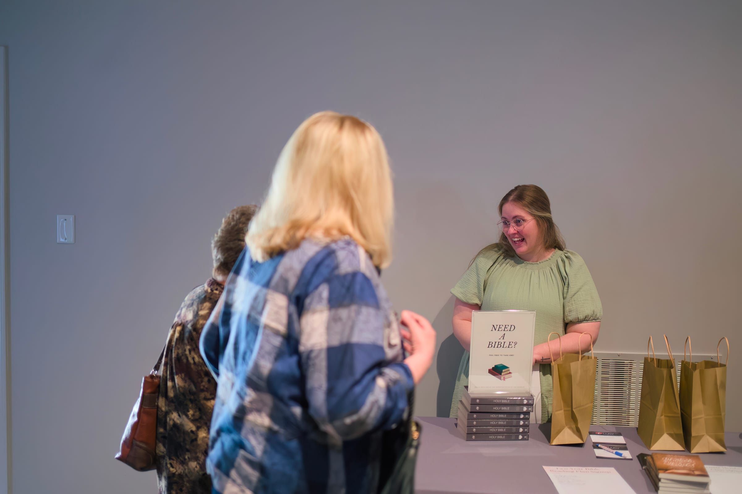 A volunteer smiles while greeting visitors at the welcome table, beside a "Need a Bible?" sign, stacked Bibles, and gift bags for guests.