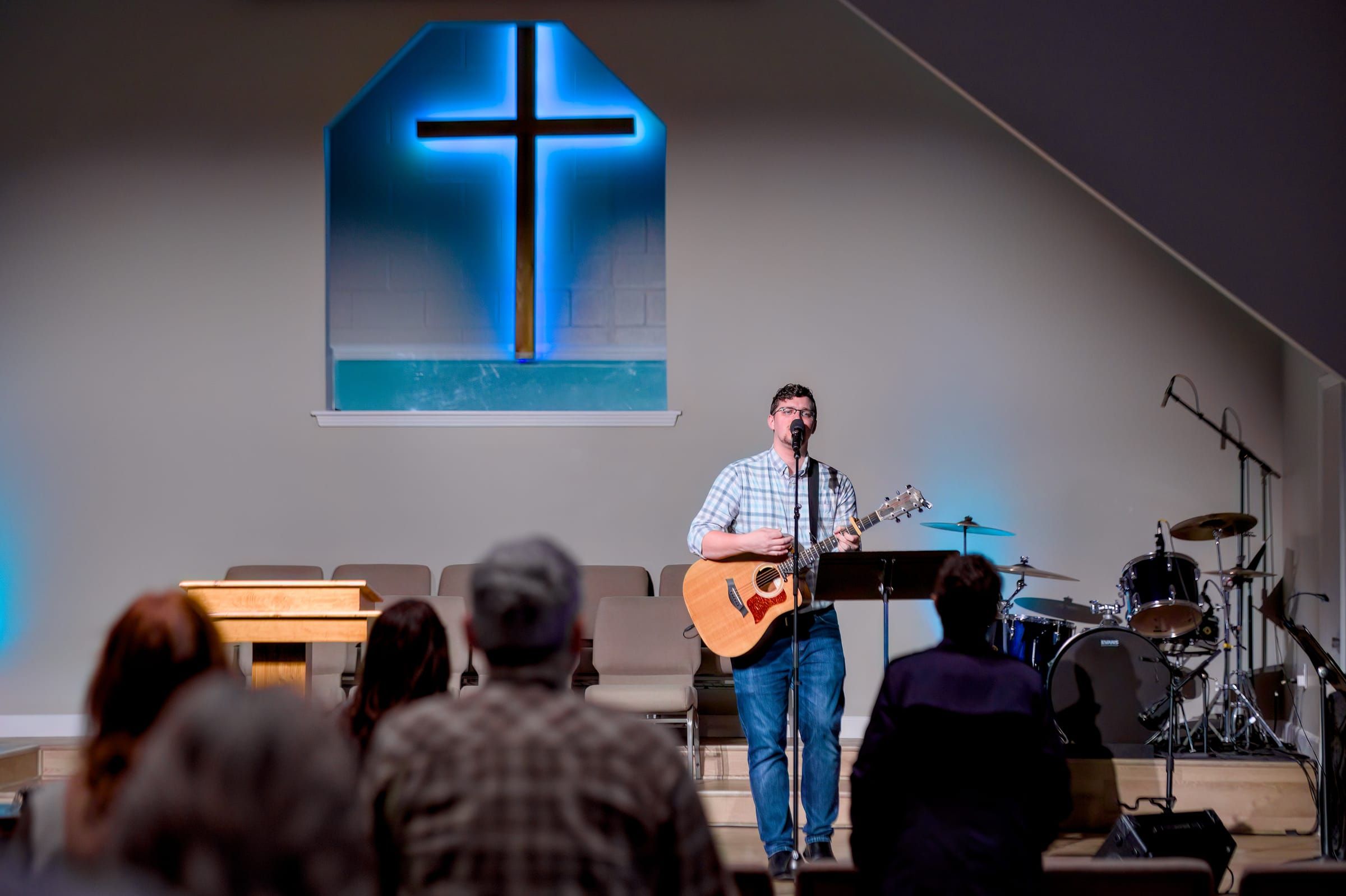 A worship leader singing and playing guitar on stage during a Sunday gathering, the congregation gathered below.