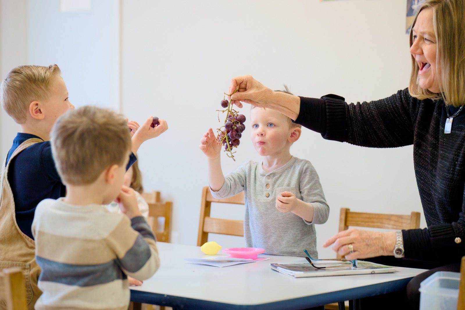 A young boy in a gray shirt reaching up for a bunch of grapes a teacher holds out during an object lesson in a kids ministry class.