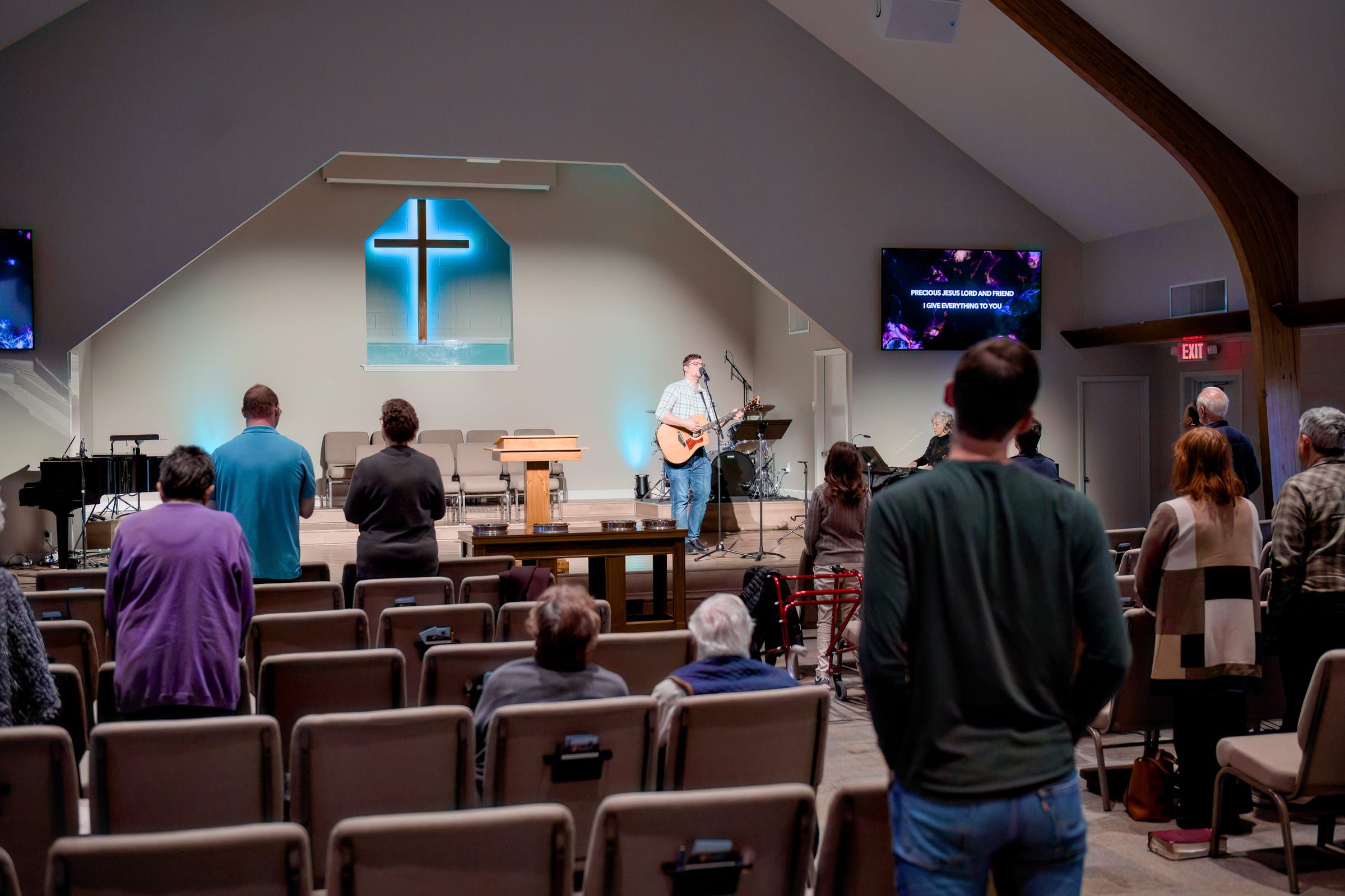 The congregation standing in worship as the band leads from the stage, an illuminated cross above them.