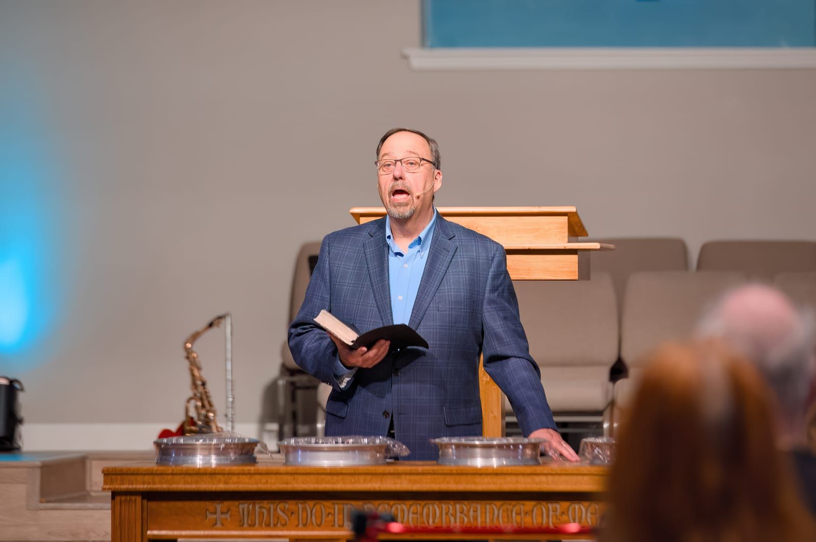 A pastor speaking at the communion table inscribed “This do in remembrance of Me,” with the congregation gathered below.