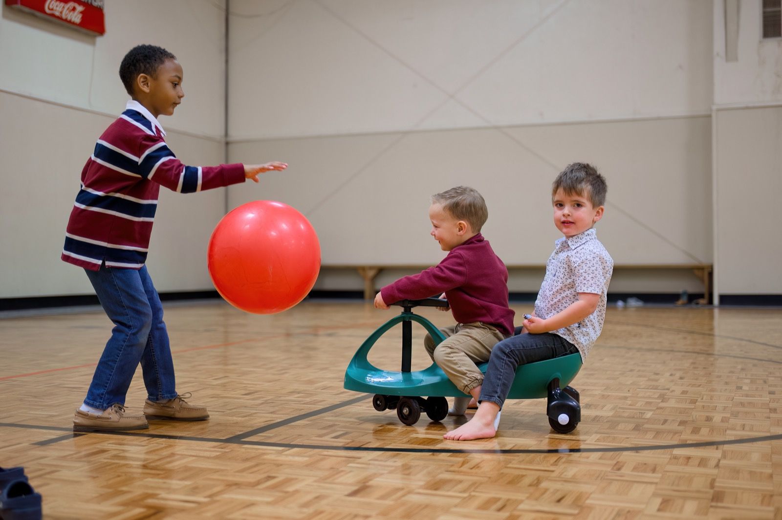 Children playing with ride-on toys and a ball in the Lucy Baptist gymnasium.