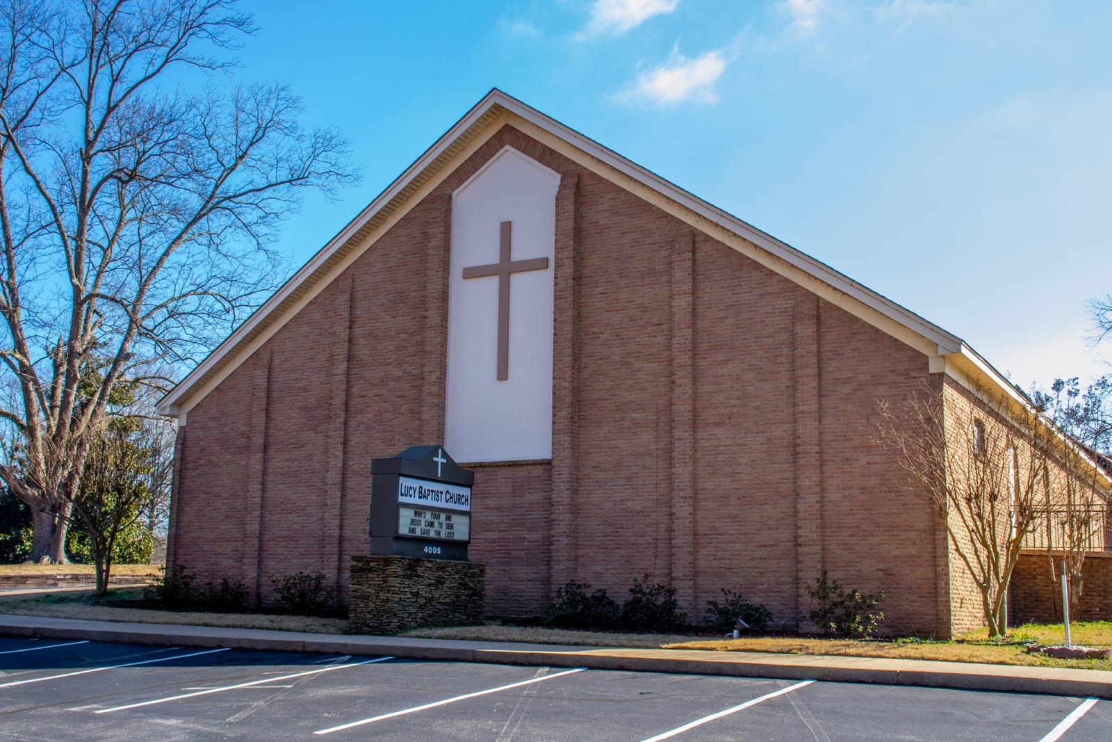 The Lucy Baptist Church building and front parking lot on a sunny day, with the church sign and cross out front.
